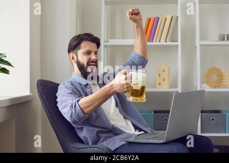 Young guy with mug of beer watching football online on laptop at home. Sports fan cheering for his soccer team indoors Stock Photo