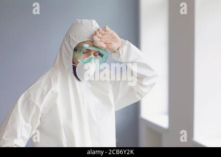 Exhausted doctor in protective hazmat suit wiping forehead at clinic. Medic working on viral epidemic prevention indoors Stock Photo