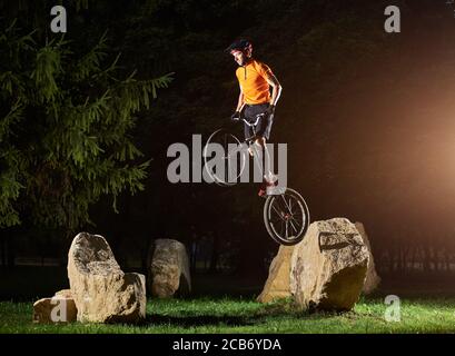 Side view of fearless guy doing trick while standing on a bike. Cyclist holding handlebar in hand and doing stunt on stone. Concept of motion.