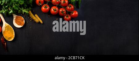 Food background with colorful food ingredients border from above. Top view of parsley, cherry tomato, herbs and spices on rustic black table. Culinary Stock Photo