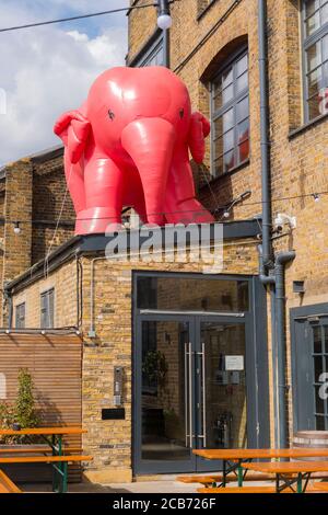 England London Stratford Park Hackney Wick giant inflatable pink elephant over entrance glass doors building tables benches drainpipes cables windows Stock Photo