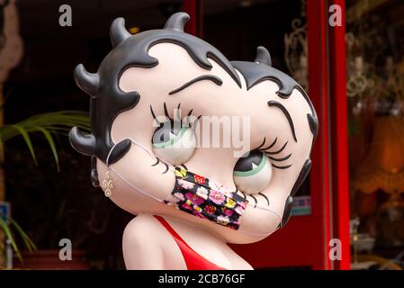 Betty Boop wearing a COVID-19 face mask. Large caricature of the Max Fleischer cartoon film star with a Coronavirus face covering. Red dress. Humorous Stock Photo