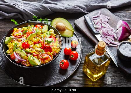 sweet corn salad with avocado, red onion slices and tomatoes in a black bowl on a dark wooden table with ingredients on dark old cutting board, mexica Stock Photo