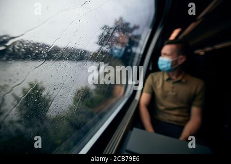 Man wearing face mask inside train in rainy day. Selective focus on raindrops on window. Stock Photo