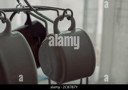 Coffee Mugs Hanging on Rack. White porcelain cups on metal hooks. Shooting at eye level. Selective focus. Stock Photo