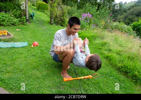 Father crouching on lawn grass in country garden playing holding balancing his 3 year old son child boy in summer garden Wales UK KATHY DEWITT Stock Photo
