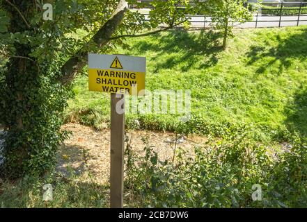 A yellow warning triangle with shallow water on it next to a small stream. Stock Photo