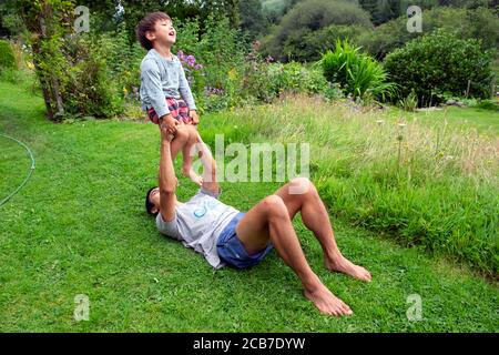 Father lying on the ground grass in country garden playing with balancing his 3 year old son child boy holding him in the air Wales UK KATHY DEWITT Stock Photo