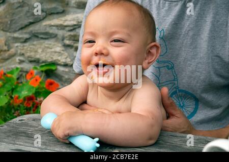 Mixed race smiling baby holding a plastic spoon sitting outside eating dinner at a picnic table being held by father in summer Wales UK  KATHY DEWITT Stock Photo