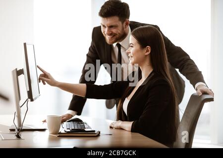 Successful business concept. Young businesswoman sits at the office table busy working on computer. Happy smiling businessman points at monitor. Two Stock Photo