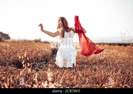 Back view of young, ginger girl in white dress running through the wheat field, holding a red scarf