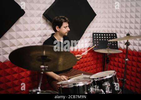 Young male drummer playing drums and cymbals. Handsome man with moustache playing with drumsticks while sitting at drums in studio. Stock Photo