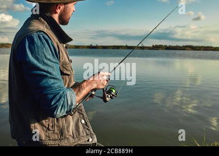 Caucasian adult bearded men stand near lake and hold fishing rod. he looks into distance and at float. Fishing is meditation. Stock Photo