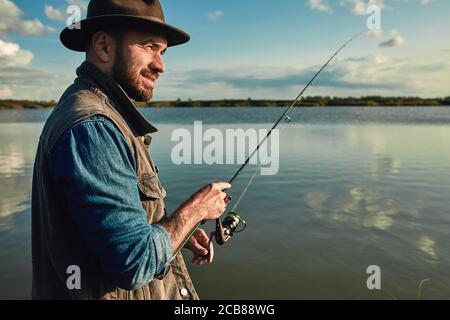 Caucasian adult bearded men stand near lake and hold fishing rod. Here he with his son, he shows how hold fishing rod correctly. Stock Photo