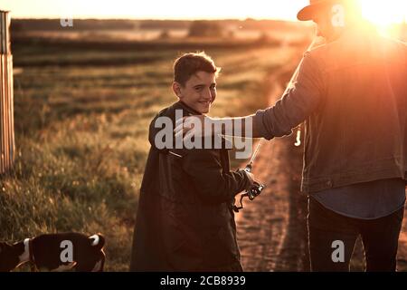 Father and son took fishing rods and fishing tackle. They go on country road near lake for fishing. Boy smile and look at camera. Sunset. Stock Photo