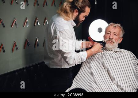 serious handsome old man sitting on the chair and having his hair, beard cut, close up portrait, man is preparing for a date. appearance Stock Photo