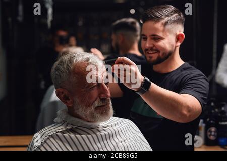 smiling awesome young barber brushing the hair of senior man, preparing to cut hair Stock Photo