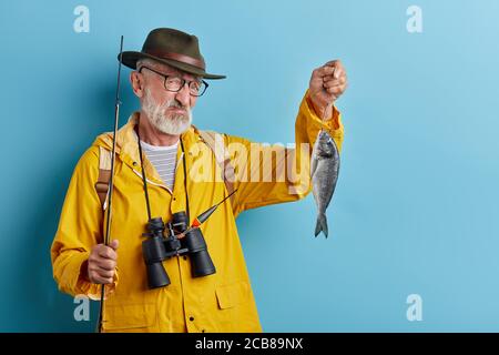 Picture of unhappy handsome old fisherman with blue eyes and beard holding fish, looking grumpy and dissatisfied, unhappy with poor catch. Hobby, leis Stock Photo