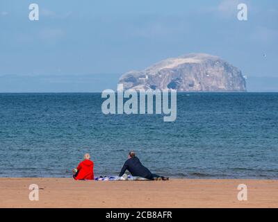 Couple sitting on beach admiring the sea view to Bass Rock gannet colony, Firth of Forth, Belhaven Bay, East Lothian, Scotland, UK Stock Photo