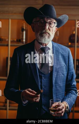 Aged old-aged dandy wearing optical glasses and big hat, dressed in dark blue elegant three piece suit smoking a cigar looking at camera with pensive Stock Photo