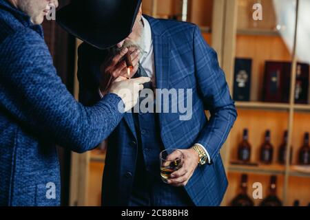 Elegant senior gentleman wearing spectacles and grey-haired beard standing at pub in wide-brimmed hat with glass of alcohol drink and cigar. Elderly m Stock Photo
