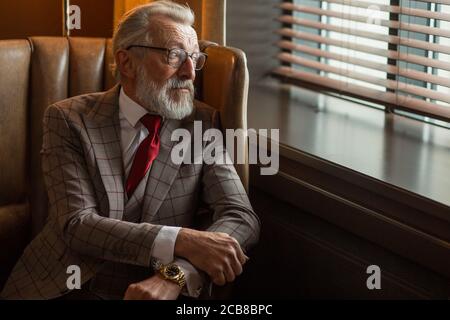 Grey haired old male business owner dressed in formal wear and spectacles, suit with red tie, posing at luxury indoor interior, looking at camera, wel Stock Photo