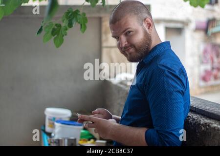 Bald bearded man wearing a blue shirt with dots leaning on a stone wall, holding a phone and looking at the camera with a serious look Stock Photo
