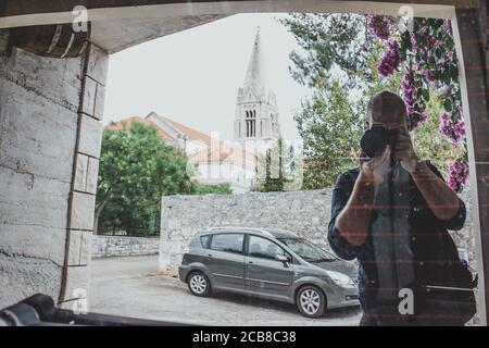 Man taking a photo of himself in the relfection of a car window, church on the island of brac in croatia visible in the reflection. Typical traveler i Stock Photo
