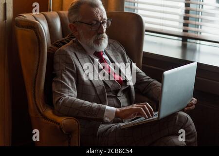 Fashionable focused elderly male writer wearing glasses and elegant clothes working on laptop, close up. Old author creating new story while sitting i Stock Photo