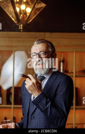 Confident elderly man in blue expensive suit clothes enjoying a rest, relaxing in private gentleman s club, sipping old whiskey and smoking cigars is Stock Photo