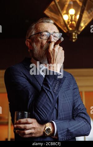 Confident elderly man in blue expensive suit clothes enjoying a rest, relaxing in private gentleman's club, sipping old whiskey and smoking cigars is Stock Photo