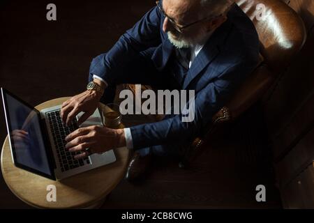 Handsome old male pensioner dressed in smart formal style using laptop and smoking while sitting on couch at luxury home, view from above. Stock Photo