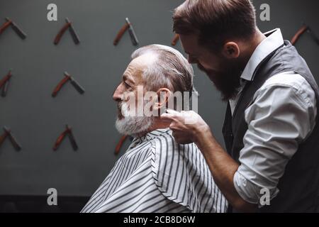 Professional young barber giving a haircut with concentration to a rich respectable gentleman, standing and holding razor while relaxed elderly client Stock Photo