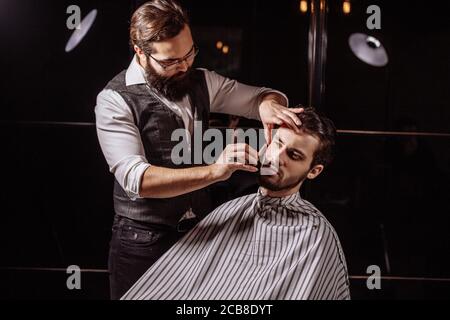 Young brunette male man s barber making an old-fashioned shave with straight razor to a young man in a stylish man s beauty salon. Stock Photo