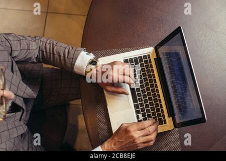 High angle view of male hands with gold wristwatch, dressed in grey expensive suit, working on laptop over red wood table background Stock Photo
