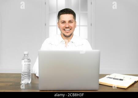 Young man smiling as he reads the screen of a laptop computer while relaxing working on a comfortable place by the wooden table at home Stock Photo