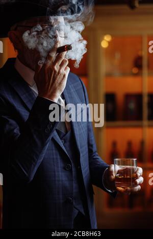 Concept of old age, wealth, prosperity and experience. Prosperious confident aged man with grey beard and spectacles, wearing big dandy hat smoking ci Stock Photo