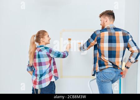 Team of two professional male and female young painters in checkered wear with brushes and rollers working indoors, making wall colour renovation of t Stock Photo