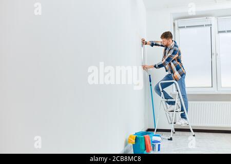 Male architect or interio designer measuring wall with flexible ruler standing on ladder in empty white appartment Stock Photo