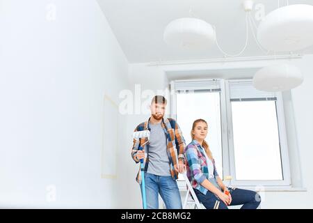 Portrait of caucasian attractive couple in checkered shirts standing , holding paint roller and brush, looking at camera. Stock Photo