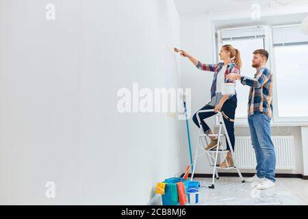 Caucasian young couple painting walls in new apartment with their own hands using paint rollers and paintbrush on wall. Stock Photo