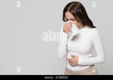 Studio portrait of female with napkin sneezing, experiences allergy symptom, isolated on grey background. Rhinitis, runny nose Stock Photo