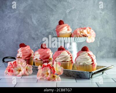 Yellow cupcakes piled high with white frosting and pink icing with a fresh raspberry on top, arranged on rustic metal trays with pink and white artifi Stock Photo