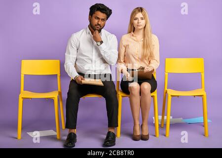 thoughtful dark-skinned man and pretty blonde woman sitting on chairs looking in camera with scattered papers all over the floor and waiting for job i Stock Photo