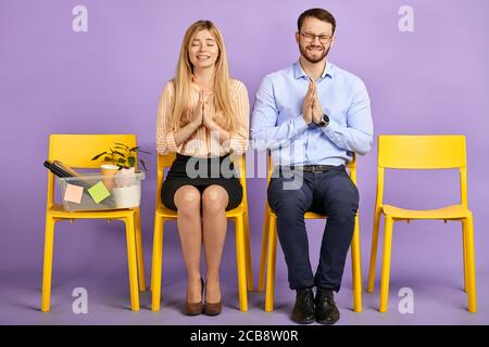 pretty young woman and man with closed eyes smiling and holding pleading hands and making a wish sitting on chairs and waiting for job interview
