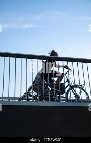 Girls cycling on bicycle bridge. Silhouettes seen from low angle through bars in metal railings. Clear blue sky with copy space. Copenhagen, Denmark - Stock Photo