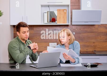 couple checking analyzing utilities bills sitting together at kitchen table, husband and beautiful wife reading bank loan documents with laptop, famil Stock Photo