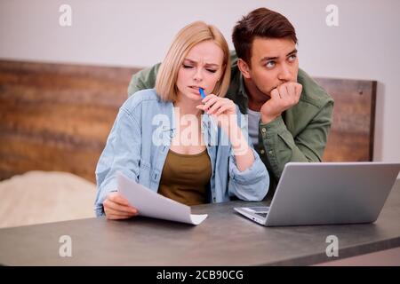 Puzzled thoughtful man and woman sitting together on table at home , studying papers or documents of their family business and working with modern lap Stock Photo