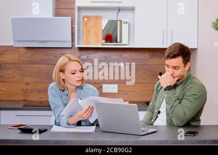 Puzzled thoughtful man and woman sitting together on table, studying documents of their family business and working with modern laptop, audit at home. Stock Photo