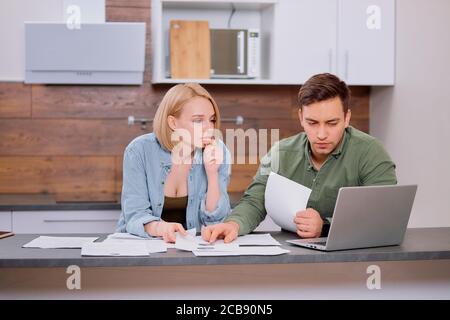 Puzzled thoughtful man and woman sitting together on table at home , studying papers or documents of their family business and working with modern lap Stock Photo